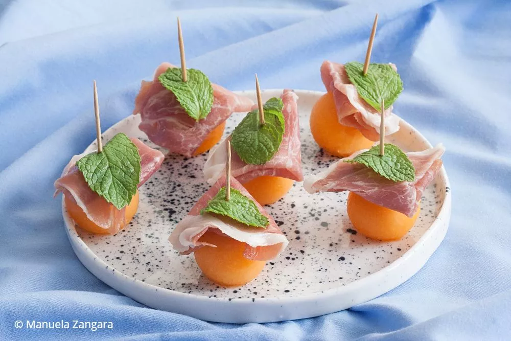 A platter of Prosciutto e Melone appetisers arranged neatly on a light blue background.