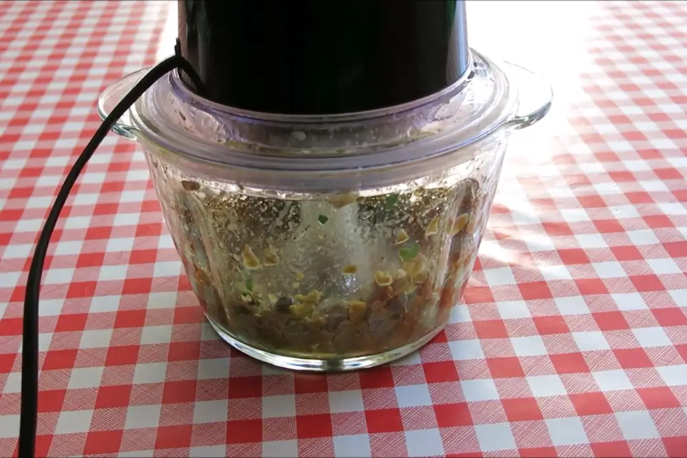 Olive mixture blending inside a glass food processor bowl with herbs and capers visible.