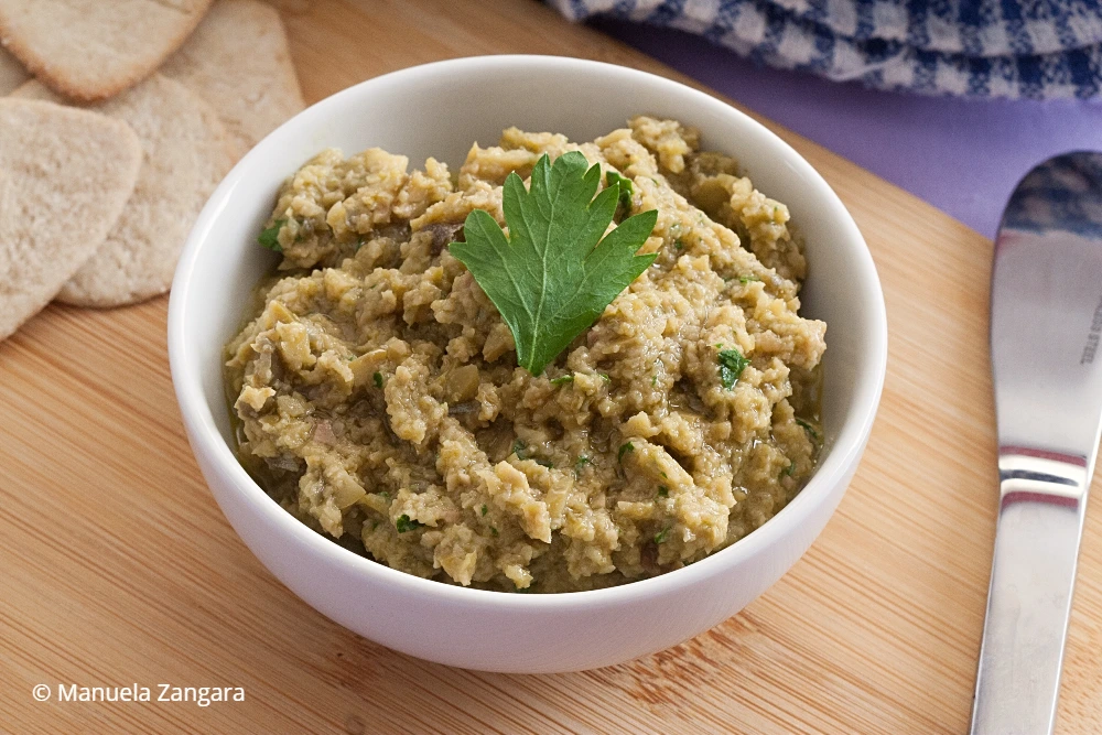 Green olive tapenade served in a white bowl garnished with fresh parsley on a wooden board.