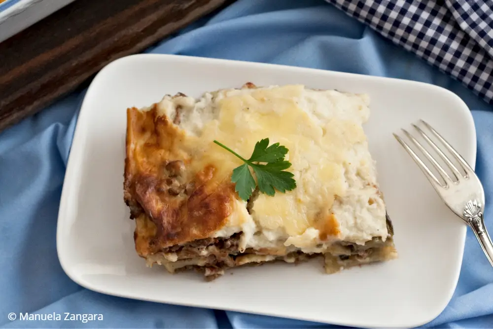 Slice of Low FODMAP Moussaka served on a white plate with creamy béchamel and visible layers.
