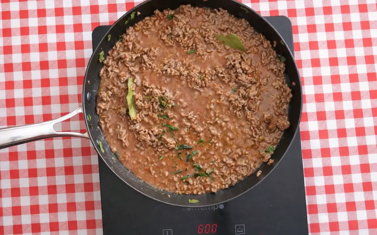 Beef mince simmering in tomato sauce with bay leaves in a frying pan.