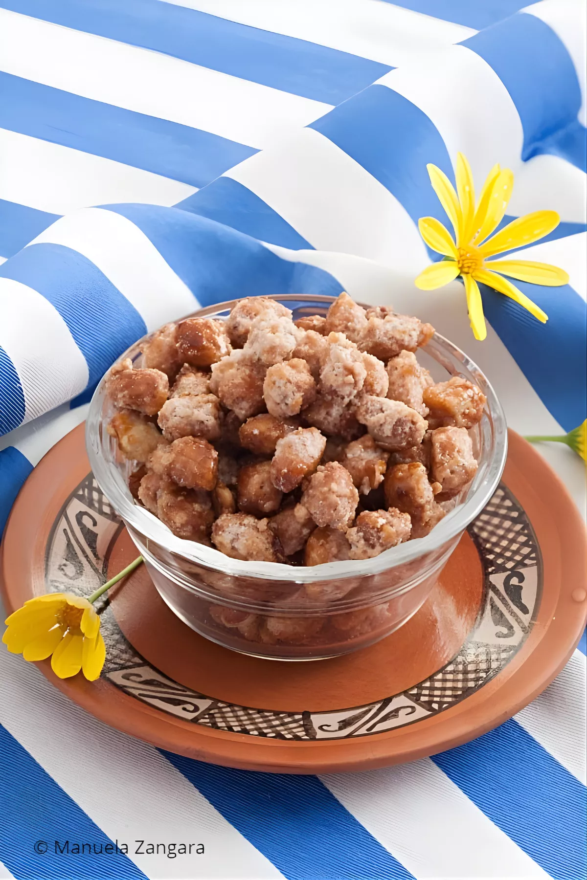 Garrapinada in a glass bowl surrounded by yellow flowers on a blue-striped background.
