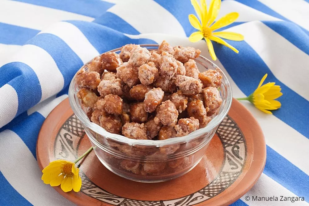 Bowl of caramelised peanuts on a terracotta plate with yellow flowers and a blue-striped cloth in the background.