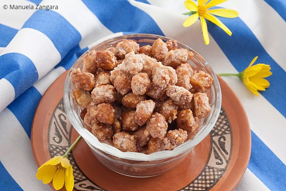 Close-up of sweet caramelised peanuts in a glass bowl with yellow flowers in the background.