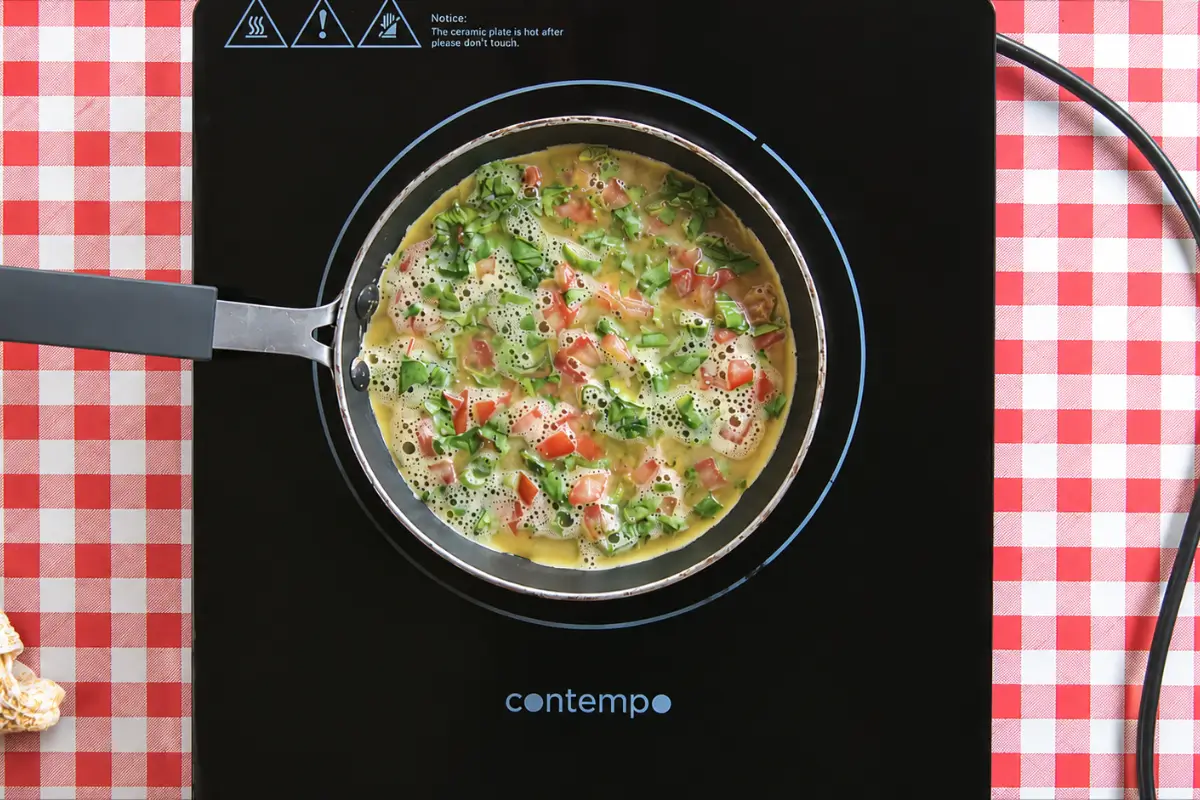 Egg mixture with tomatoes, coriander, and spring onion greens cooking in a frying pan.