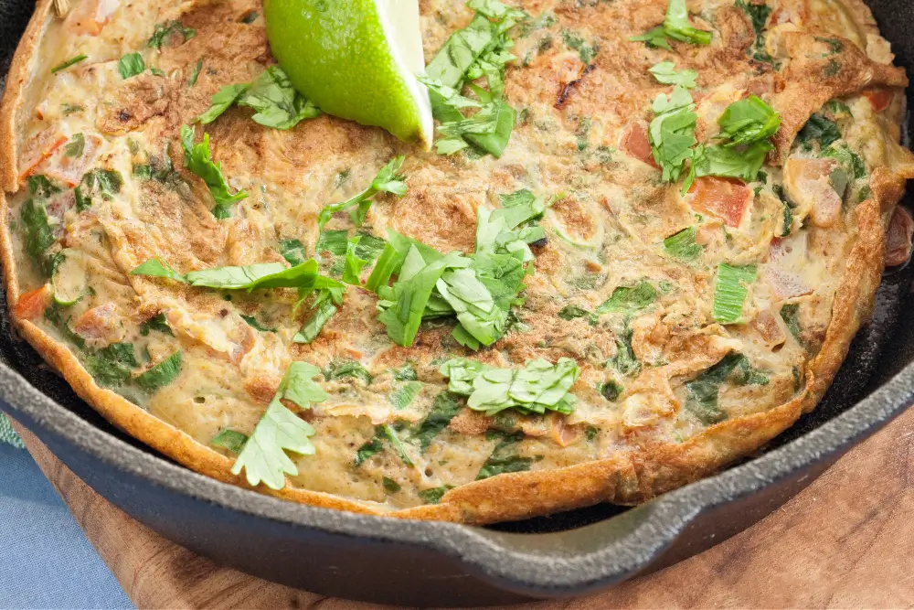 Close-up of a golden masala omelette topped with fresh coriander and a lime wedge in a cast iron pan.
