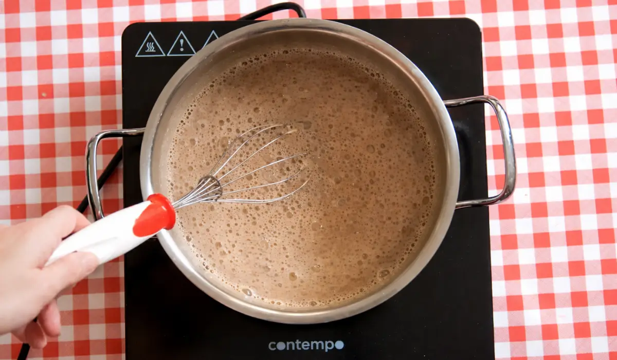 Frothy chocolate custard cooking in a saucepan while being whisked.