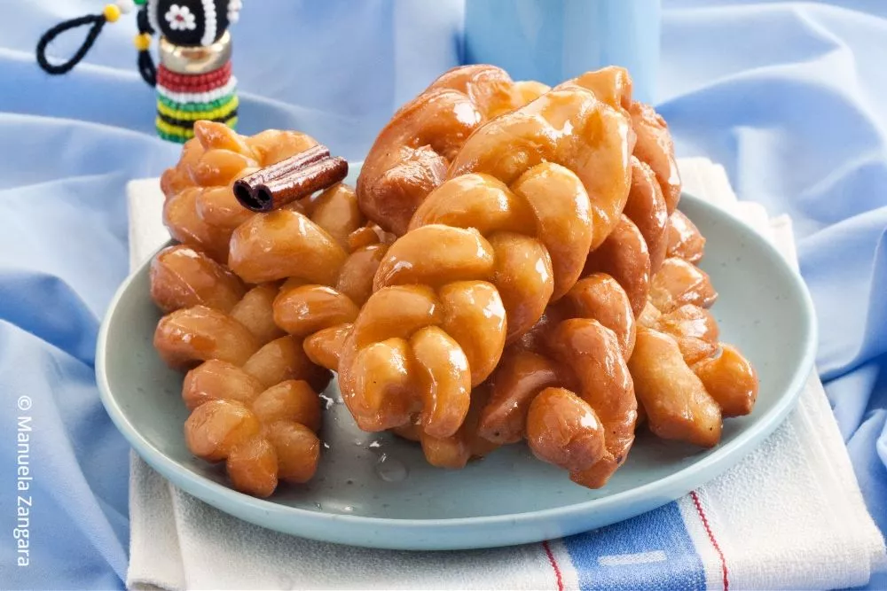 Close-up of glazed koeksisters stacked on a blue plate with glossy syrup coating.