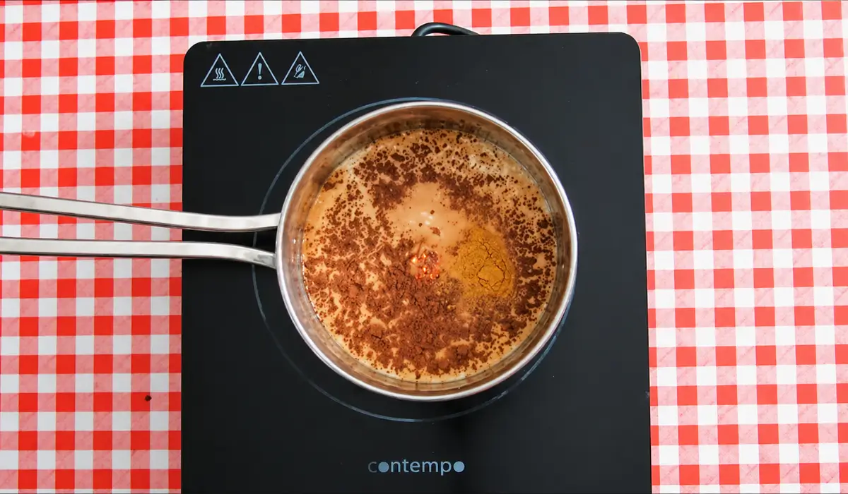 Milk heating with cocoa powder, cinnamon, and chilli flakes in a saucepan on a stovetop.