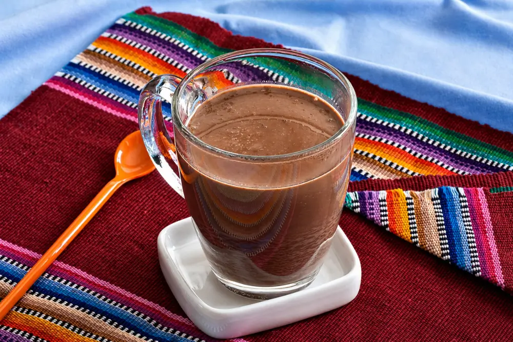 Hot chocolate served in a clear glass mug on a colourful cloth with a spoon on the side.