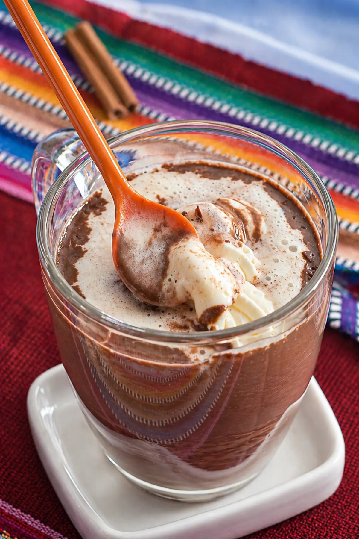 Spoon lifting whipped cream from spiced hot chocolate with a creamy surface in a glass mug.