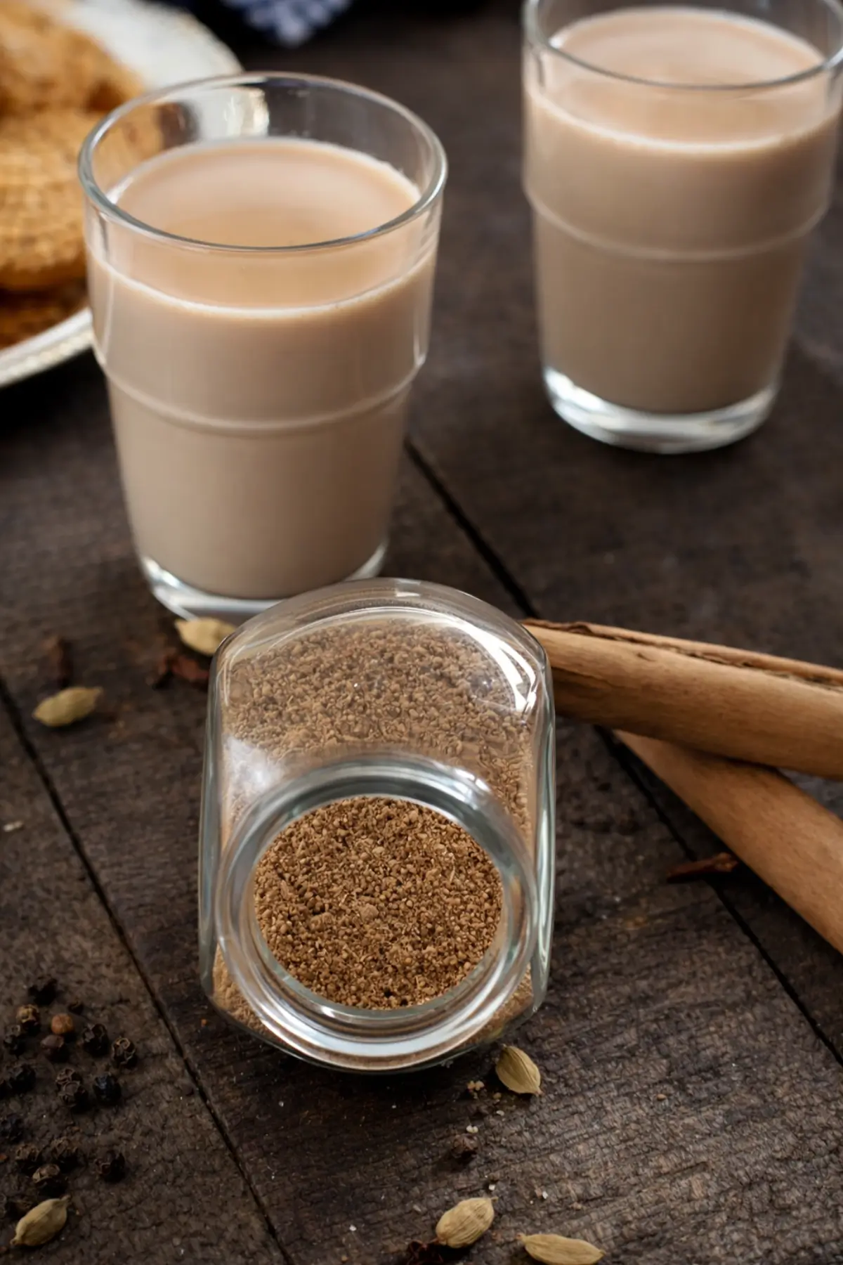 Chai masala spice blend beside two glasses of milky tea with cinnamon sticks.