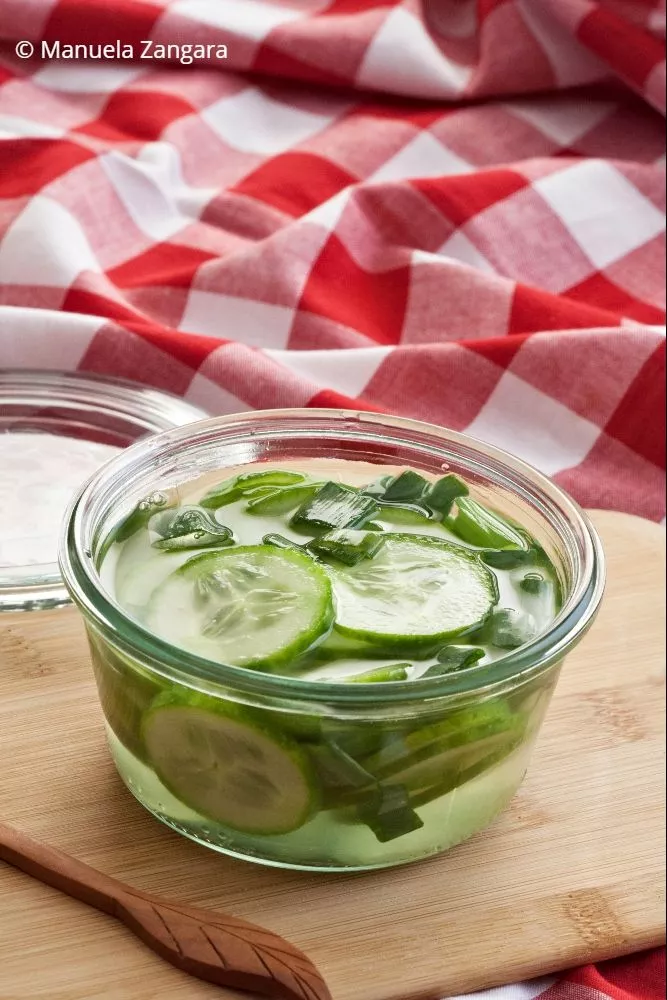 Freshly made pickled cucumbers with green onions in a clear glass jar on a wooden board.