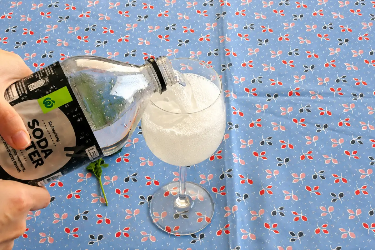 Soda water being poured into a sparkling elderflower spritz with ice in a wine glass.