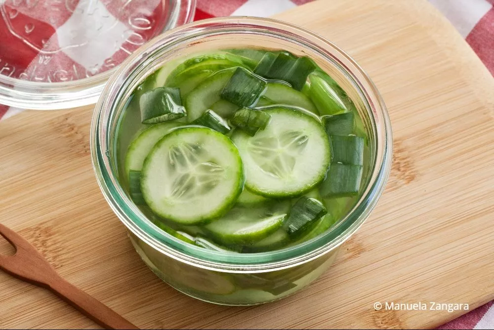 Homemade pickled cucumbers in a jar, placed on a wooden board with a red checkered cloth in the background.