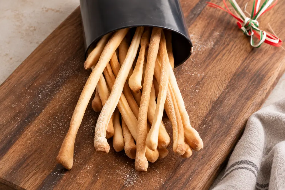 Golden Italian breadsticks spilling from a black container onto a wooden board.