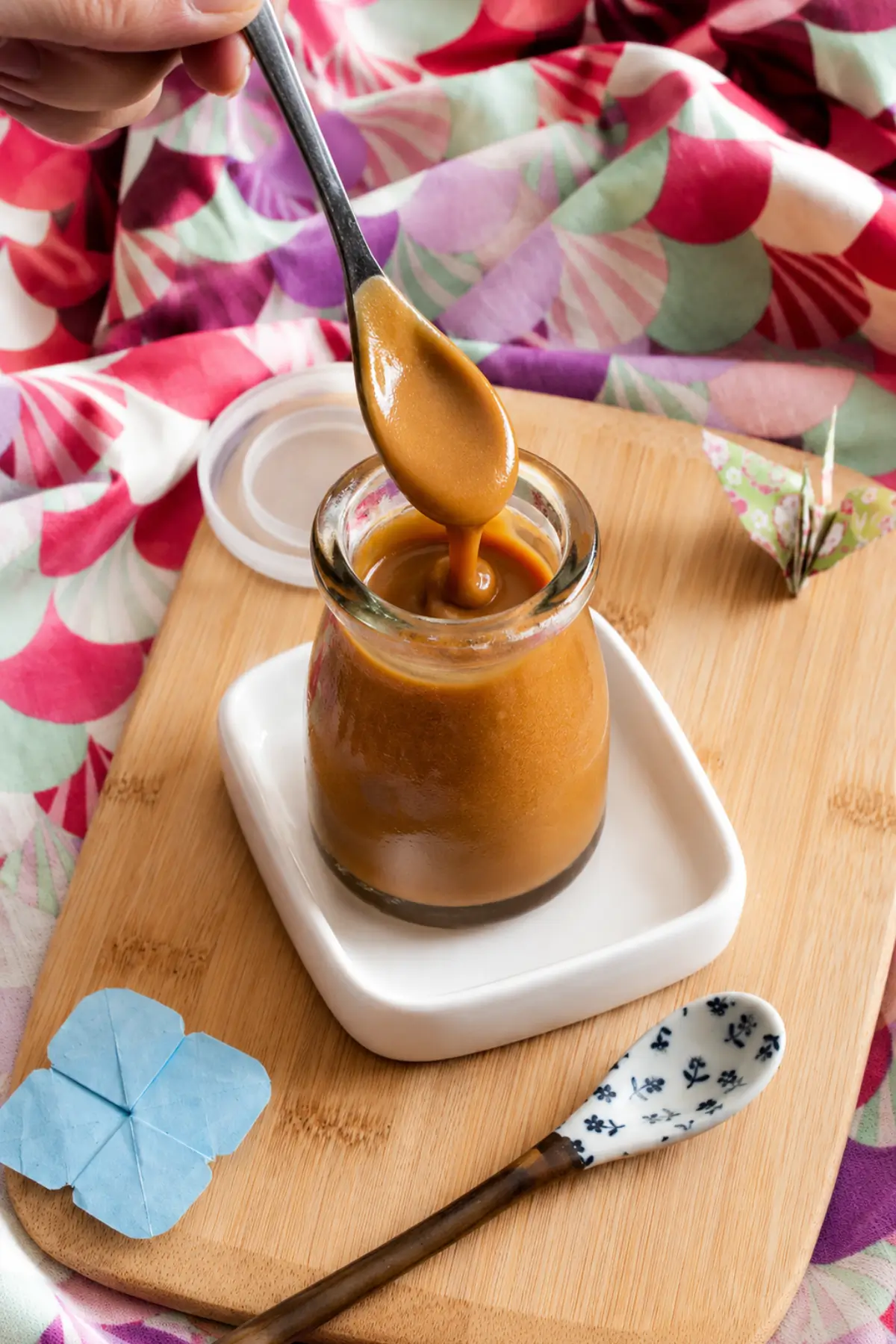 Spoon lifting thick Miso Caramel Sauce from a glass jar.