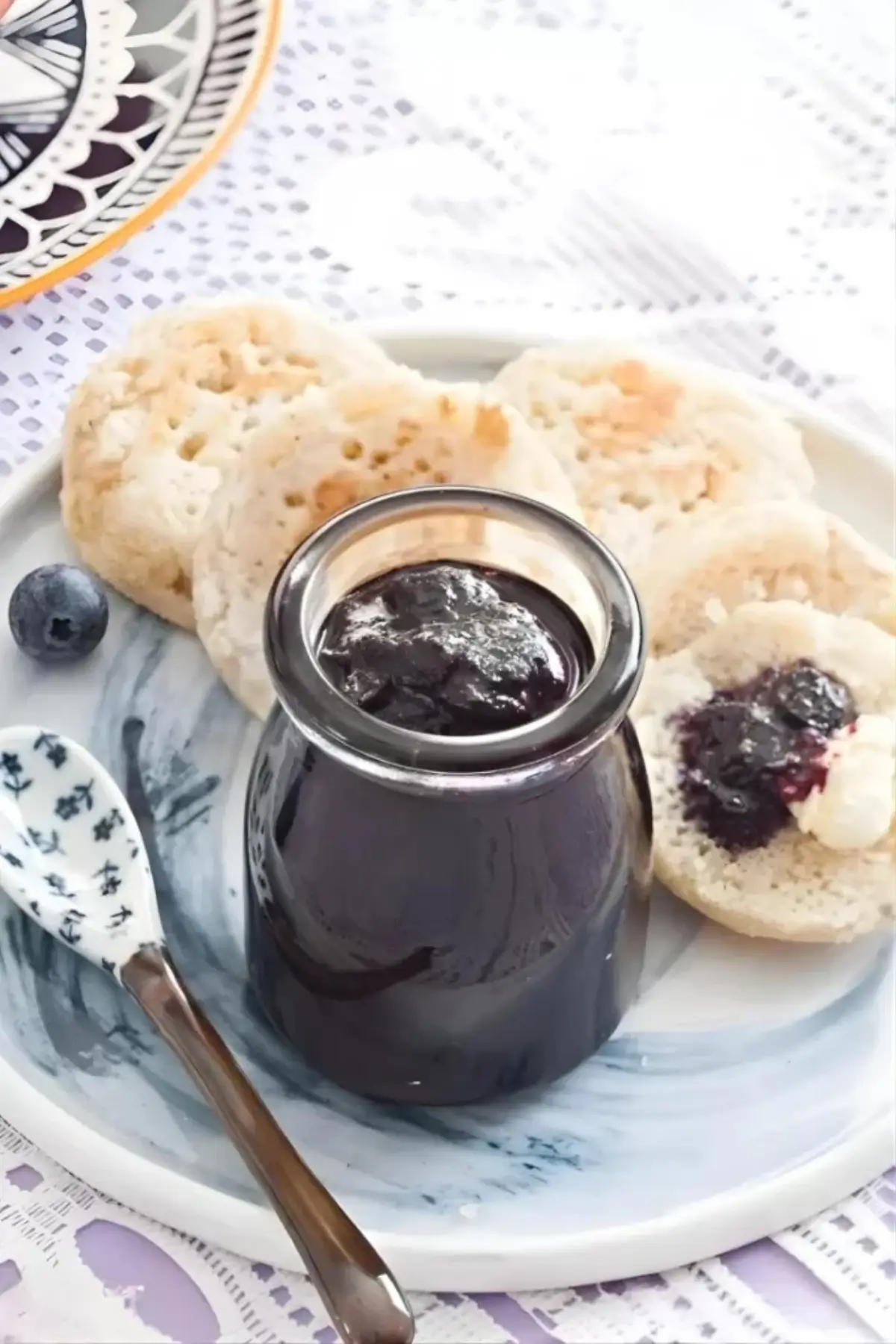 Jar of homemade blueberry jam served with crumpets, a blueberry, and a spoon on a breakfast plate.