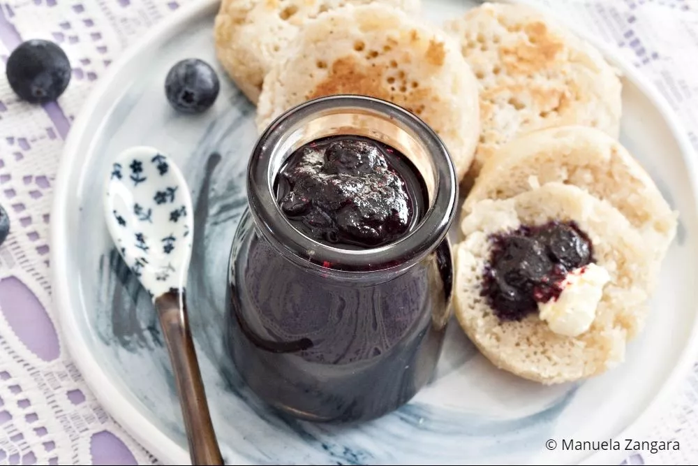 Easy blueberry jam in a small glass jar with crumpets and fresh blueberries.
