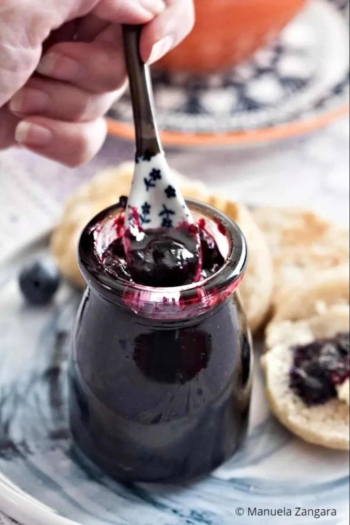 Spoon lifting a glossy scoop of homemade blueberry jam from a small glass jar.
