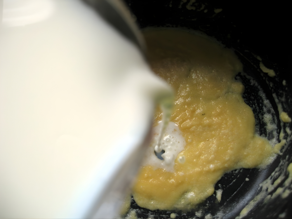 Hot milk being poured slowly into the roux while whisking to create a smooth sauce.