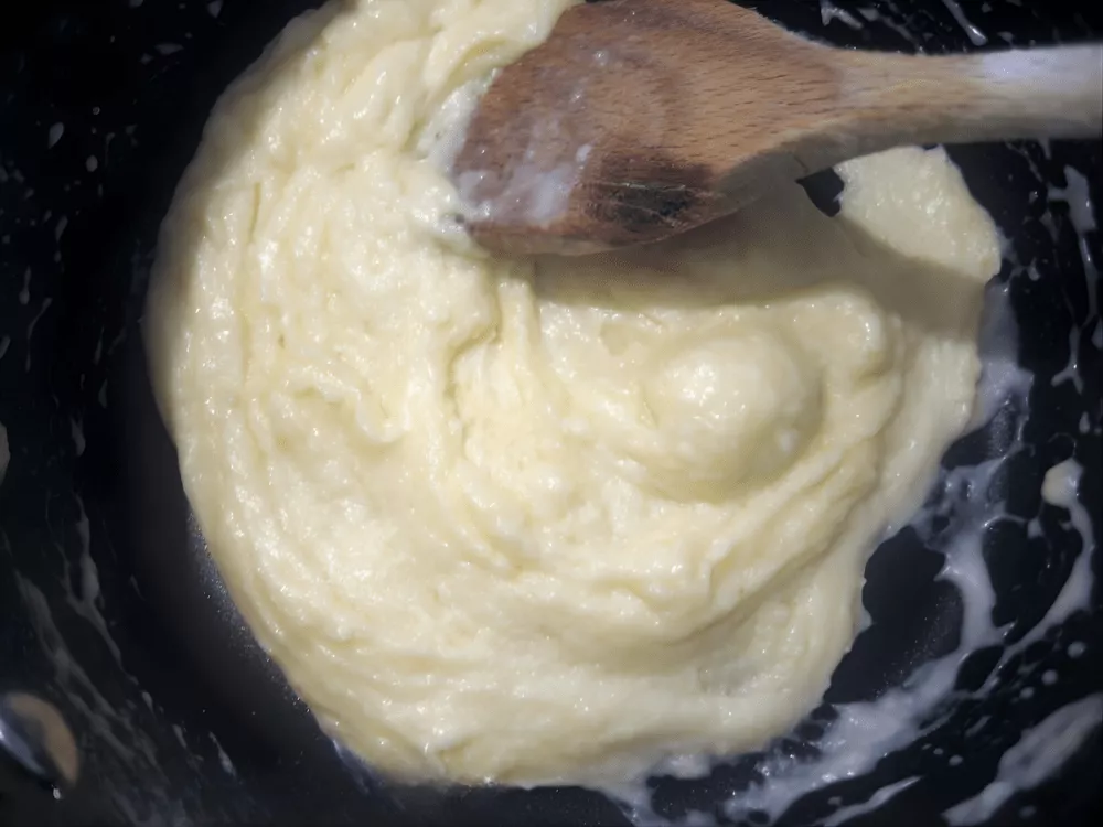Thick béchamel sauce cooking gently in a saucepan, stirred with a wooden spoon until smooth and creamy.