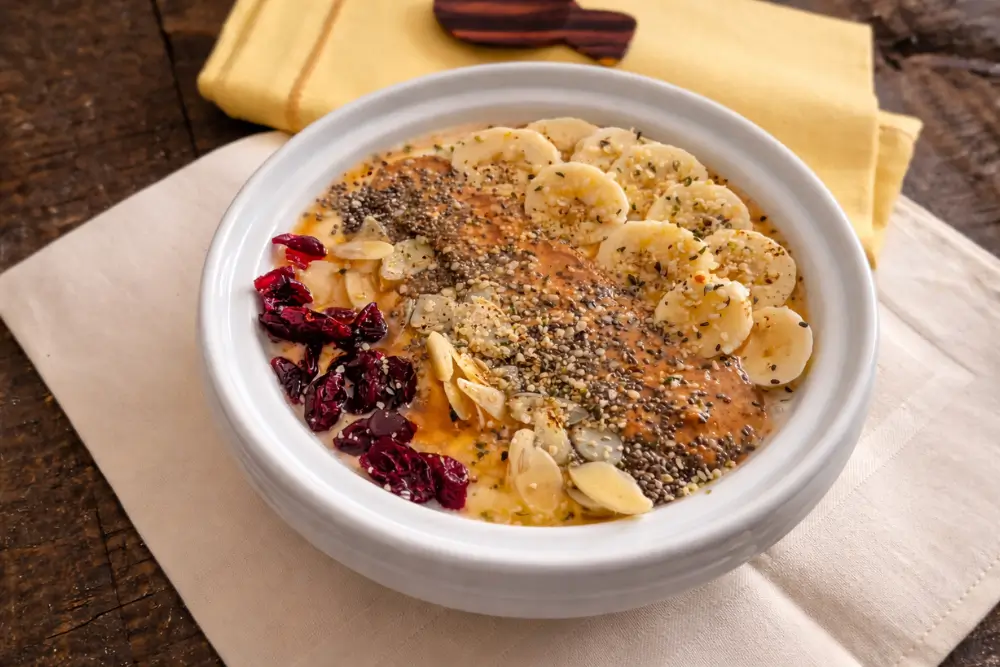 Yoghurt bowl topped with banana slices, almond butter, seeds, sliced almonds and dried cranberries on a wooden table.