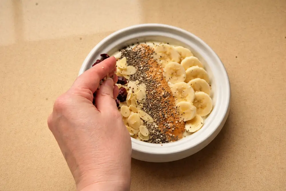 Hand sprinkling seeds over a Banana Nut Greek Yogurt Bowl topped with banana and almond butter.