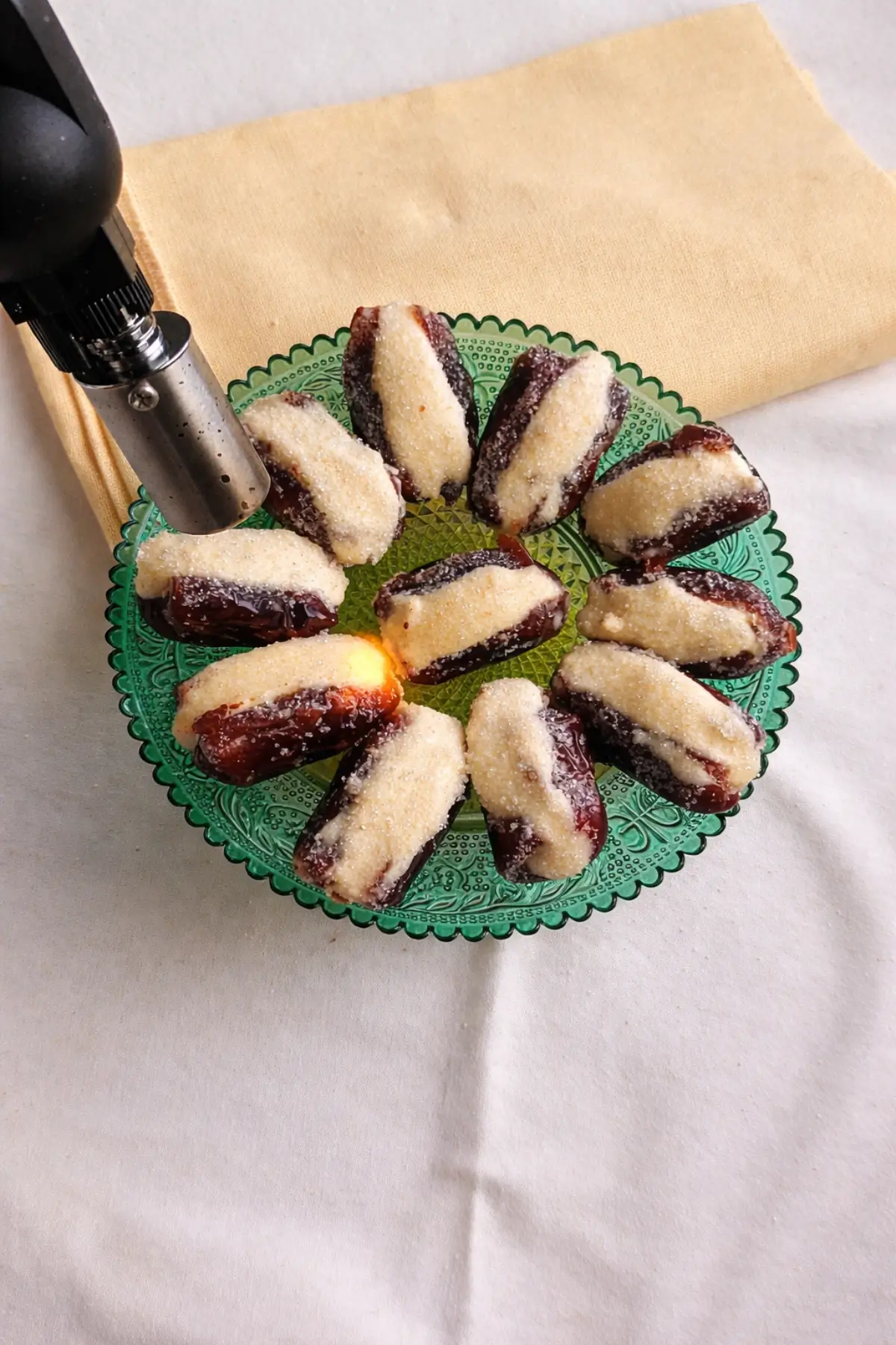 Kitchen torch caramelising sugar on stuffed dates arranged on a green plate.