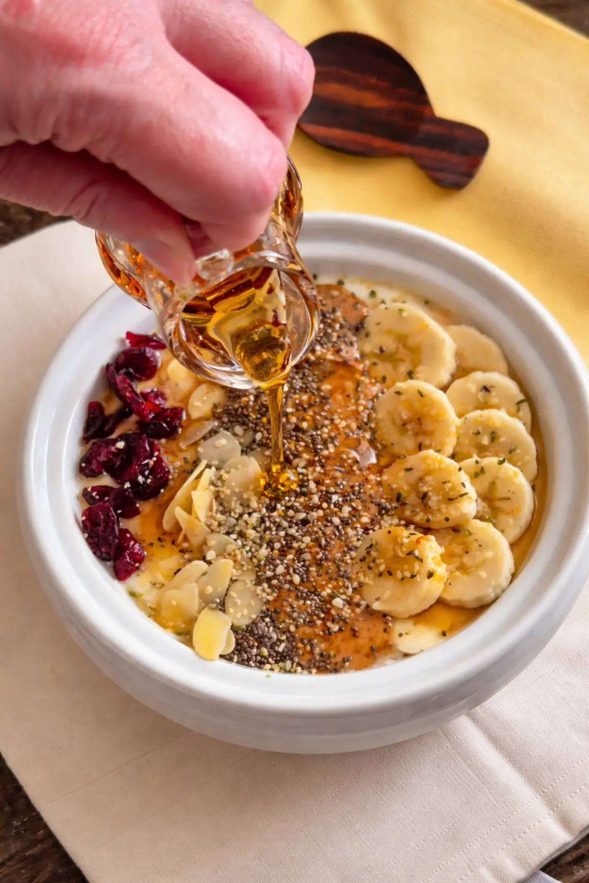 Maple syrup being poured over yoghurt topped with banana slices, seeds and nuts.