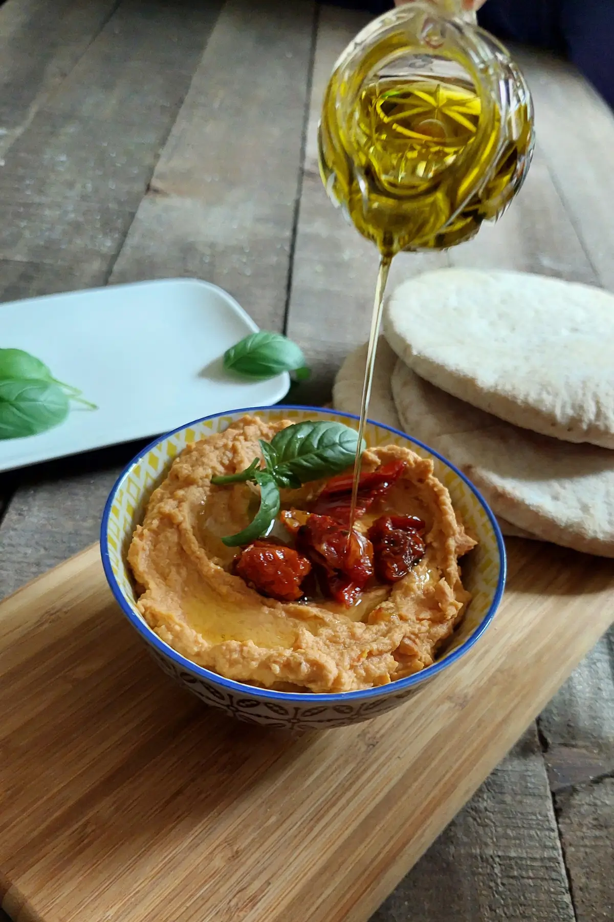 Olive oil being poured over creamy hummus topped with roasted tomatoes and basil.