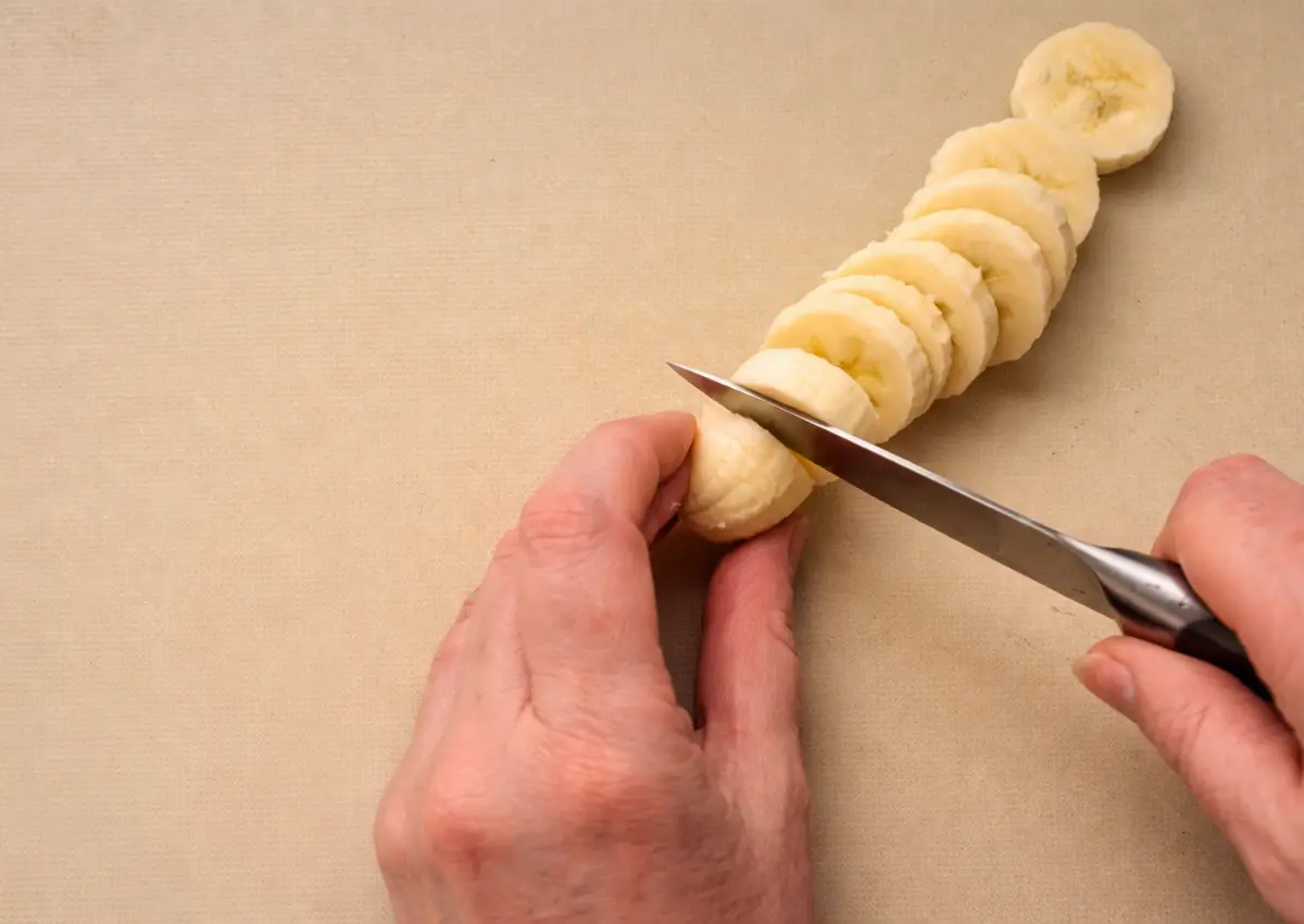 Banana being sliced into even rounds on a cutting board.