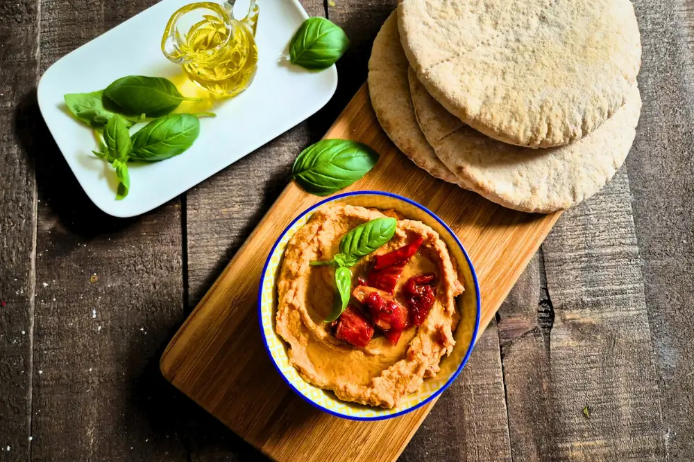 Sun-Dried Tomato Hummus served with warm pita bread and fresh basil on a wooden board.