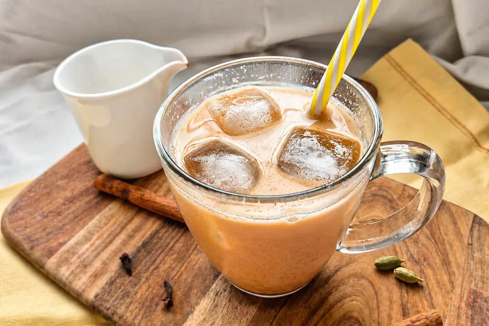 Glass of iced chai latte with milk and ice cubes on a wooden board.