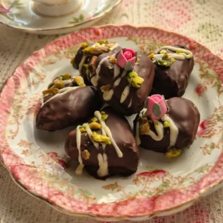 Date Chocolate Hearts decorated with white chocolate drizzle, pistachios, and sugar flowers on a vintage plate.