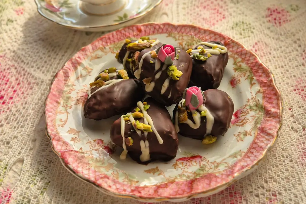 Date Chocolate Hearts decorated with white chocolate drizzle, pistachios, and sugar flowers on a vintage plate.