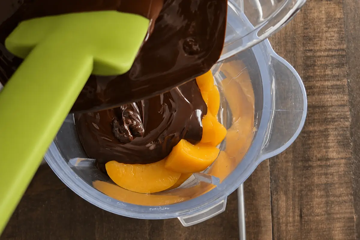 Melted dark chocolate being poured over peaches in a blender.