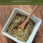 Mixed dried herbs in a white bowl with a wooden spoon resting on top.