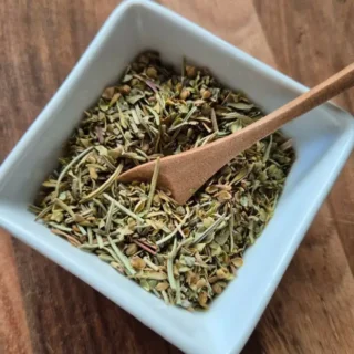 Herbes de Provence in a white bowl with a small wooden spoon on a wooden board.