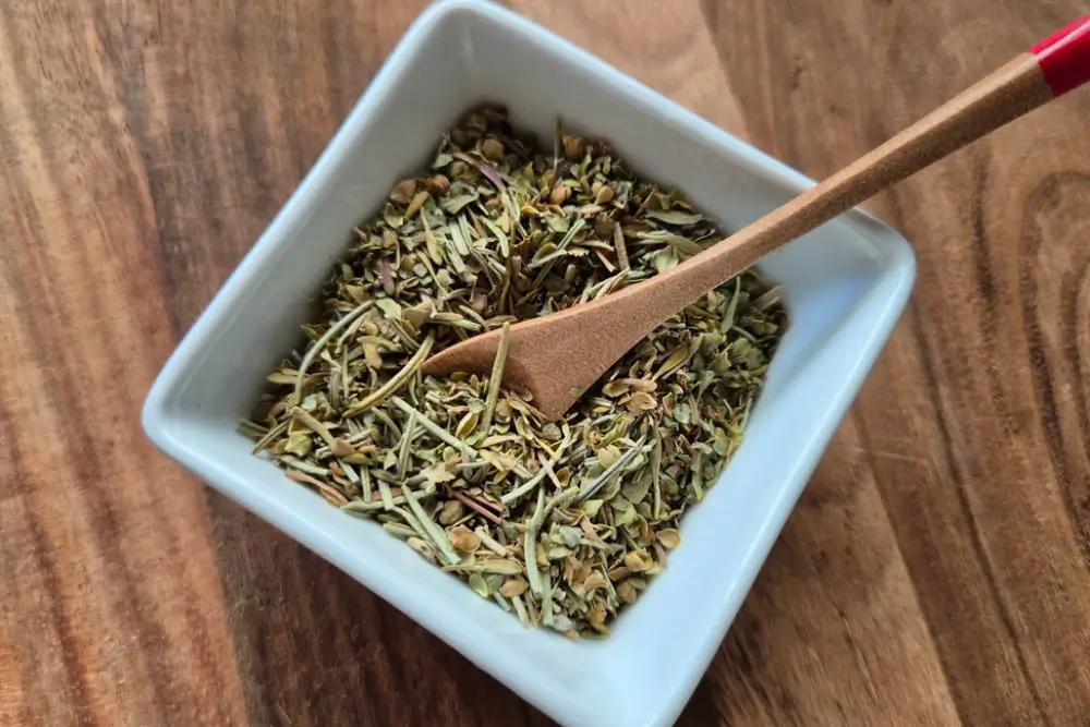 Herbes de Provence in a white bowl with a small wooden spoon on a wooden board.