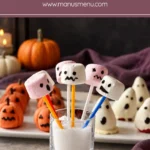 Marshmallow Monsters with pink and white faces on colourful sticks in a glass, with pumpkin and ghost treats on a platter behind.