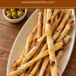 Sun-Dried Tomato Grissini piled diagonally on a speckled oval ceramic platter over a wooden table.