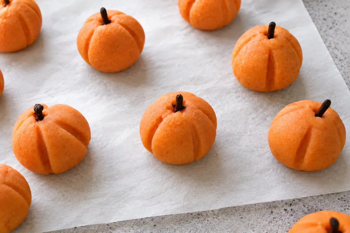 Orange marzipan pumpkins with vertical ridges and whole clove stems arranged on baking paper.