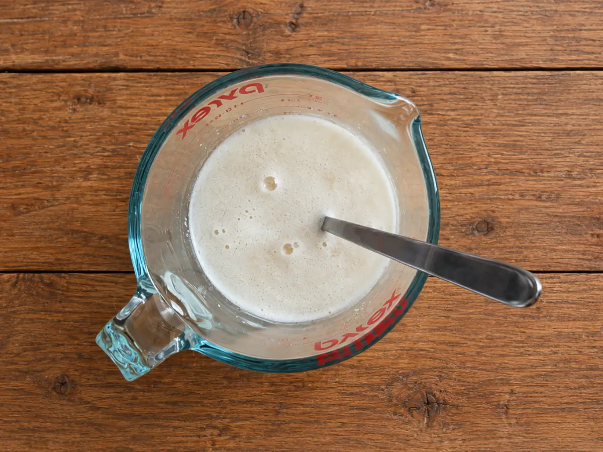 Frothy yeast mixture in a glass measuring jug with a metal spoon on a wooden table.